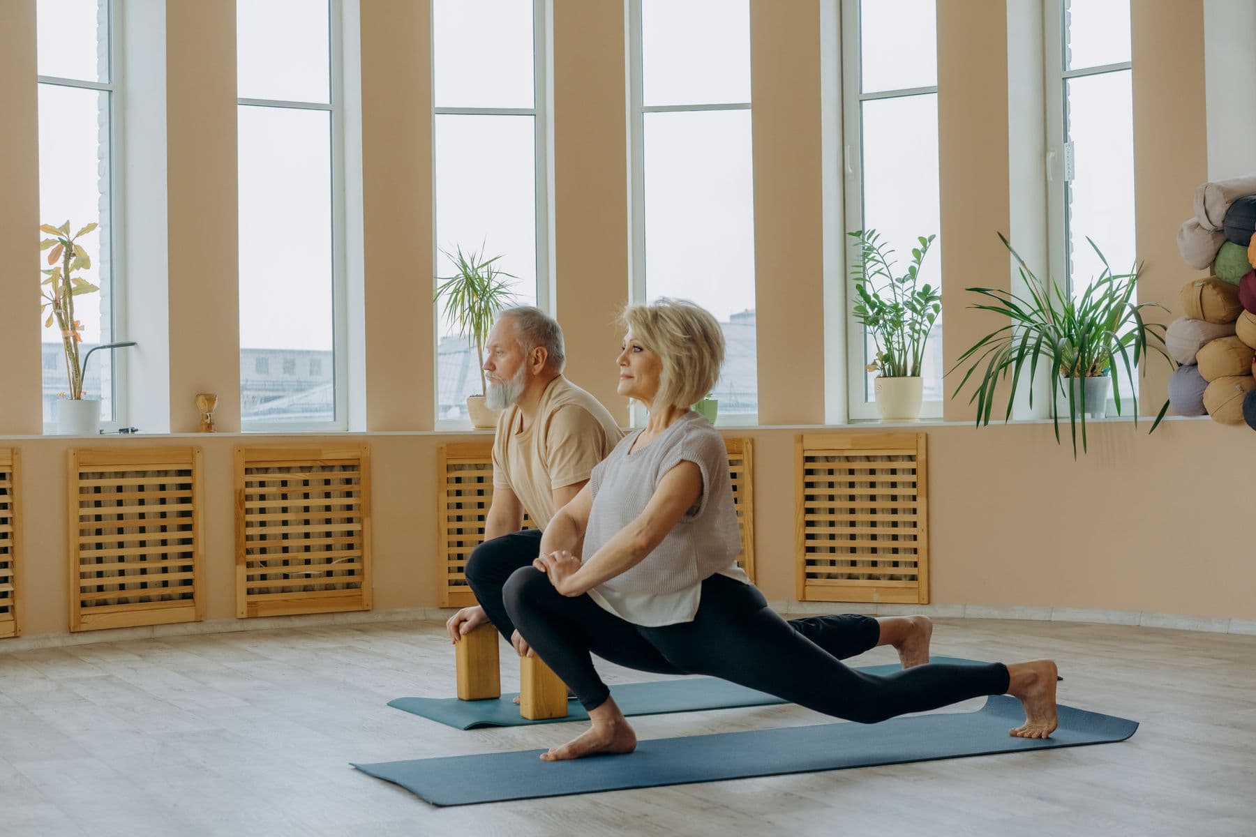 An older man and woman doing a deep lunge stretch on mats in a bright studio with tall windows.
