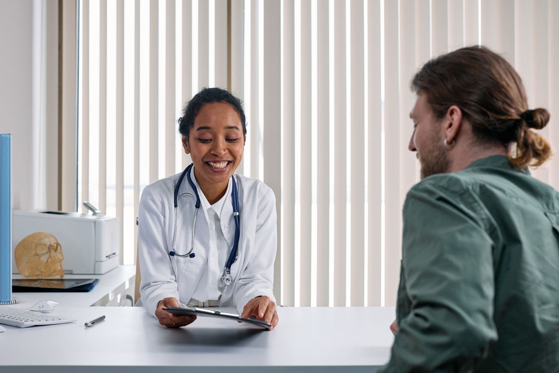 A smiling physician in a white coat, stethoscope around her neck, going through results on a tablet with a patient across her desk.