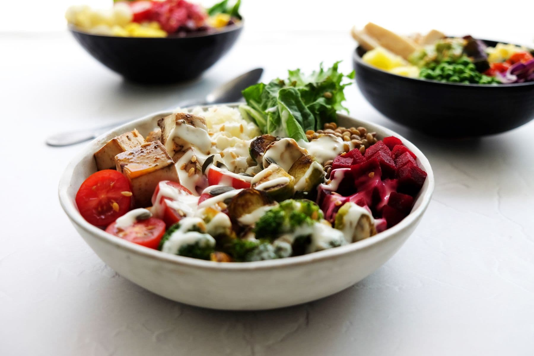 A white bowl of roasted vegetables, tofu, beetroot and greens, with more bowls behind.