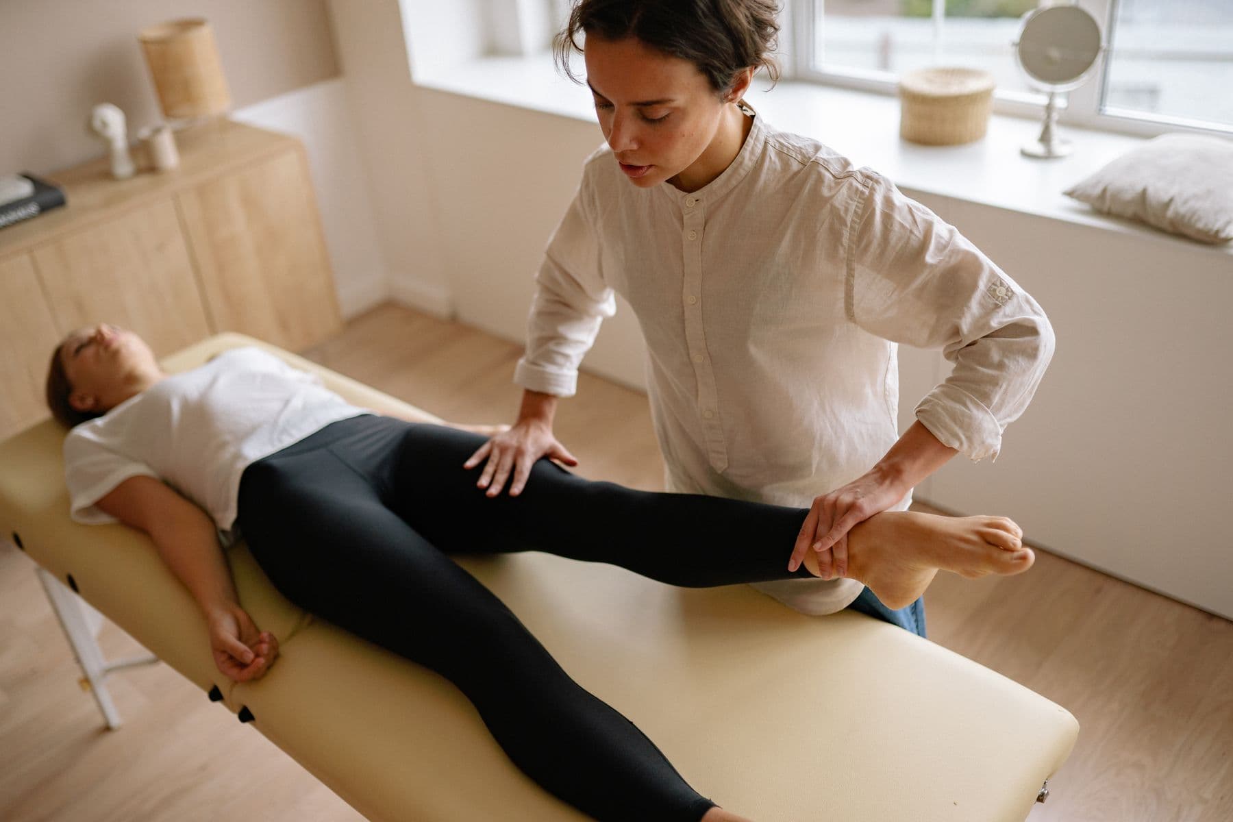 A therapist mobilising a patient's leg on a treatment couch in a light-filled room.
