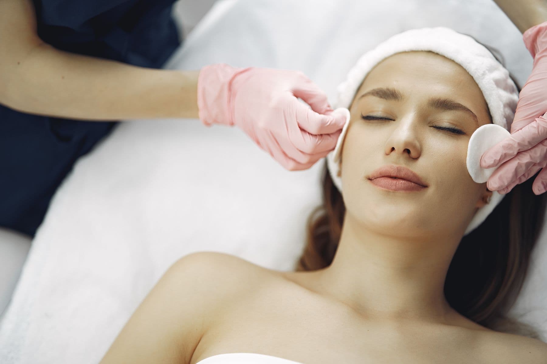 A woman with her eyes closed receiving a gentle facial cleanse from a practitioner in pink gloves.