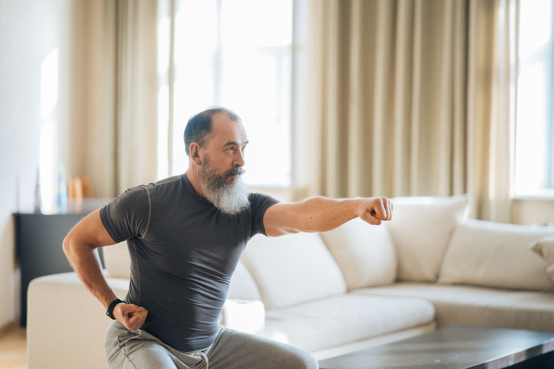A grey-bearded man in a T-shirt doing a lunge-and-punch exercise in a bright living room.